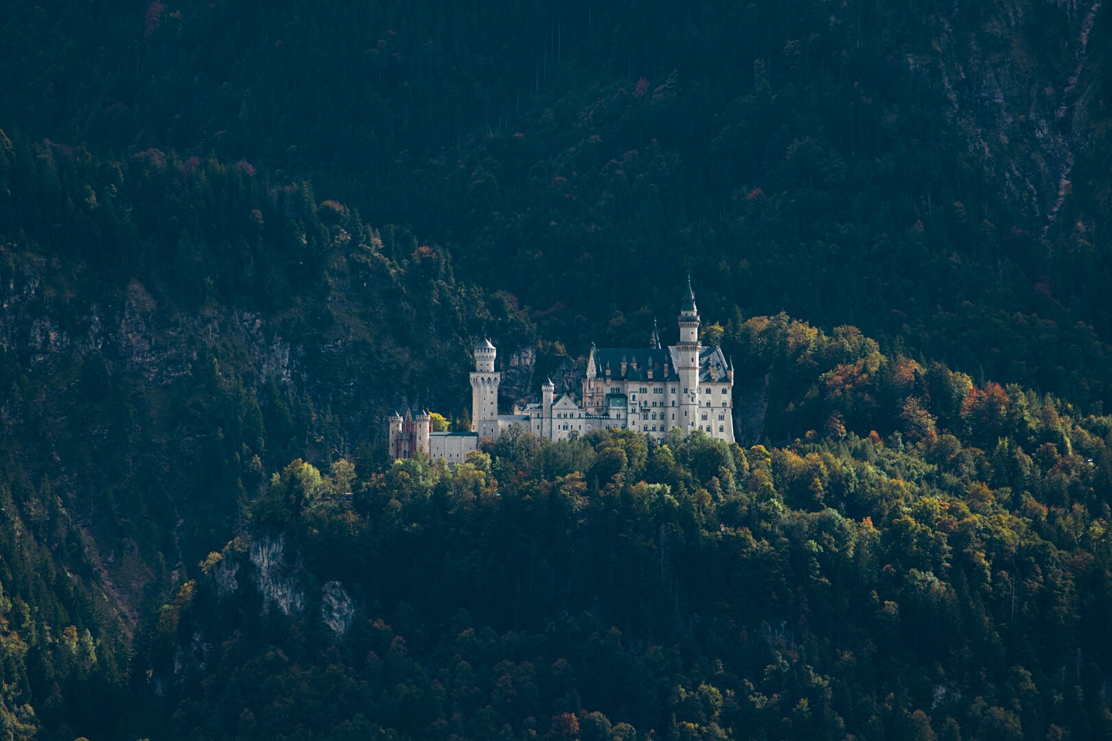 DE: Foto von Schloss Neuschwanstein umsäumt von herbstlichen Bäumen. Man erkennt links das rote Eingangstor mit zwei kleinen weißen Türmen. Weiter rechts erkennt man den weißen Hauptteil des Schlosses mit mehreren Türmen und blauen Dächern.

EN: Photo of Neuschwanstein Castle surrounded by autumnal trees. On the left you can see the red entrance gate with two small white towers. Further to the right you can see the white main part of the castle with several towers and blue roofs.