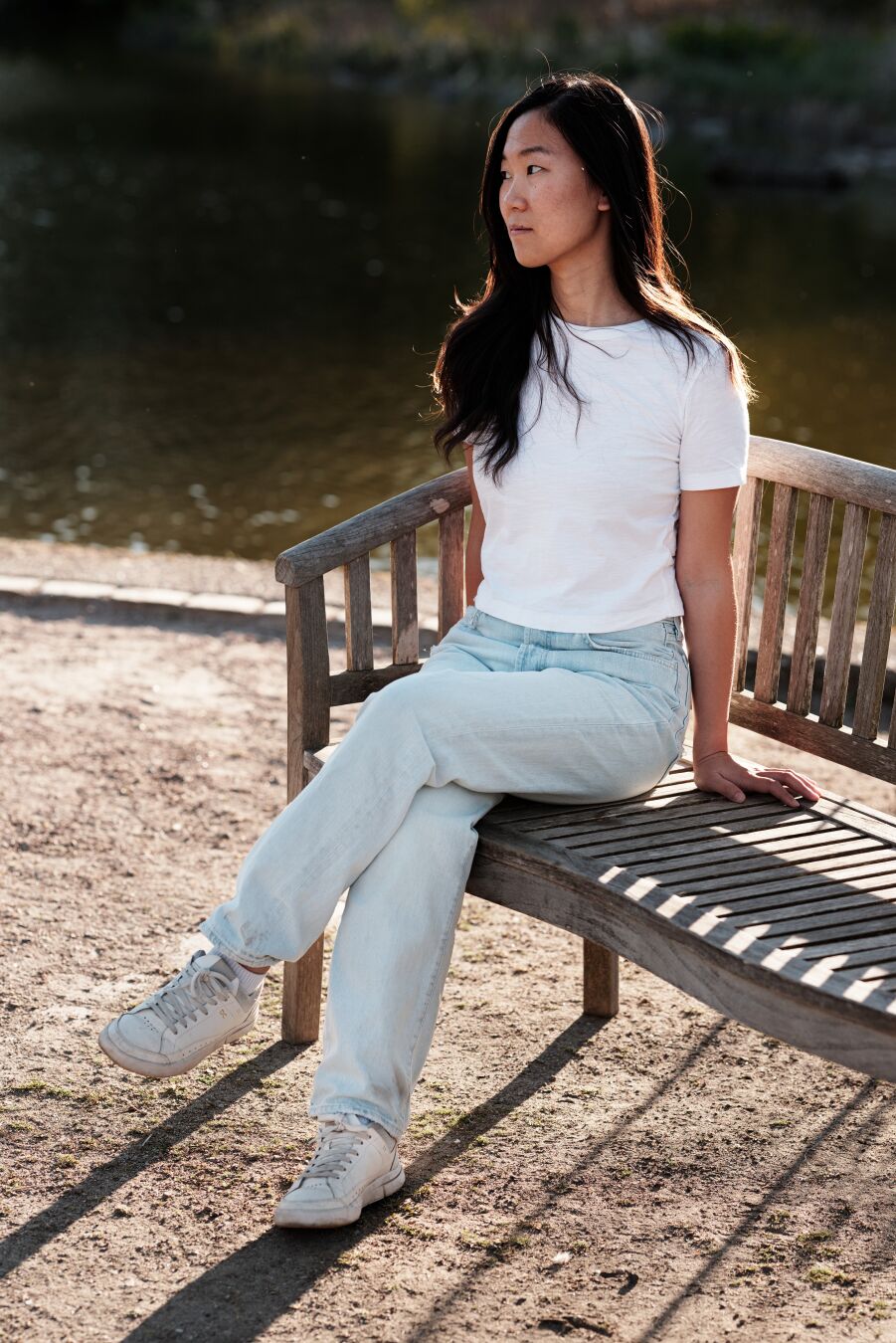 DE: Foto einer dunkelhaarigen asiatischen Frau, die auf einer Bank sitzt und nach links schaut. Sie trägt eine helle Jeans und ein weißes T-Shirt. Das Licht fällt von hinten und lässt ihr Haar leuchten. Der Bereich um die Bank herum besteht aus Erde, und dahinter ist eine Wasserfläche zu sehen.

EN: Photo of a dark-haired Asian woman sitting on a bench looking to the left side. She is wearing light-colored jeans and a white t-shirt. The light comes from behind and makes her hair glow. The area around the bench is soil, and behind it a water surface can be seen.