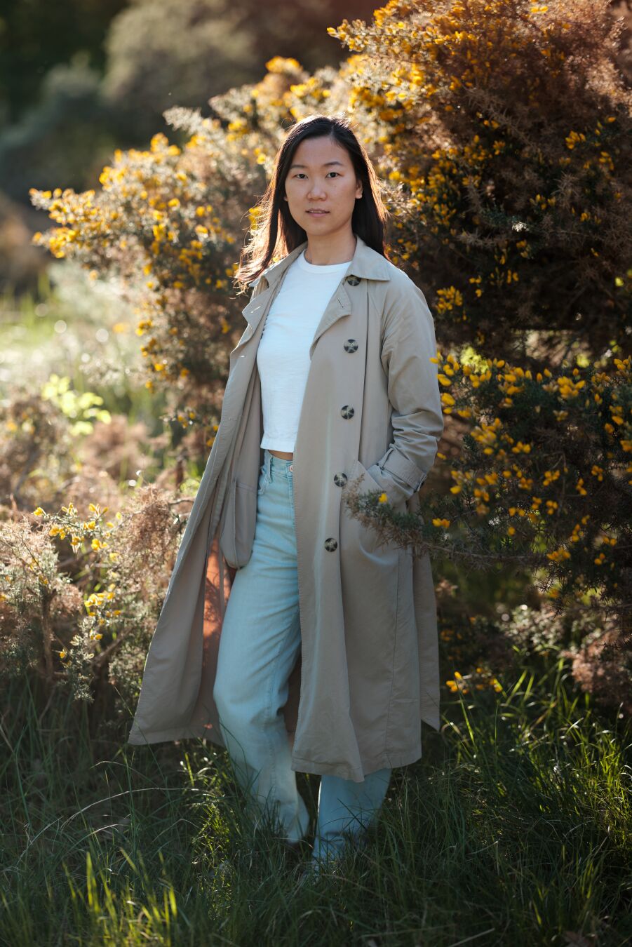 DE: Foto einer dunkelhaarigen asiatischen Frau in einem Trenchcoat, einem weißen T-Shirt und hellen Jeans. Sie steht vor einem gelb blühenden Baum. Das Licht fällt von hinten und lässt ihr Haar leuchten. Im Hintergrund sind grüne und gelbe Pflanzen zu sehen.

EN: Photo of a dark-haired Asian woman in a trench coat, white t-shirt, and light-colored jeans. She is standing in front of a yellow blooming tree. The light comes from behind and makes her hair glow. The background consists of green and yellow plants.
