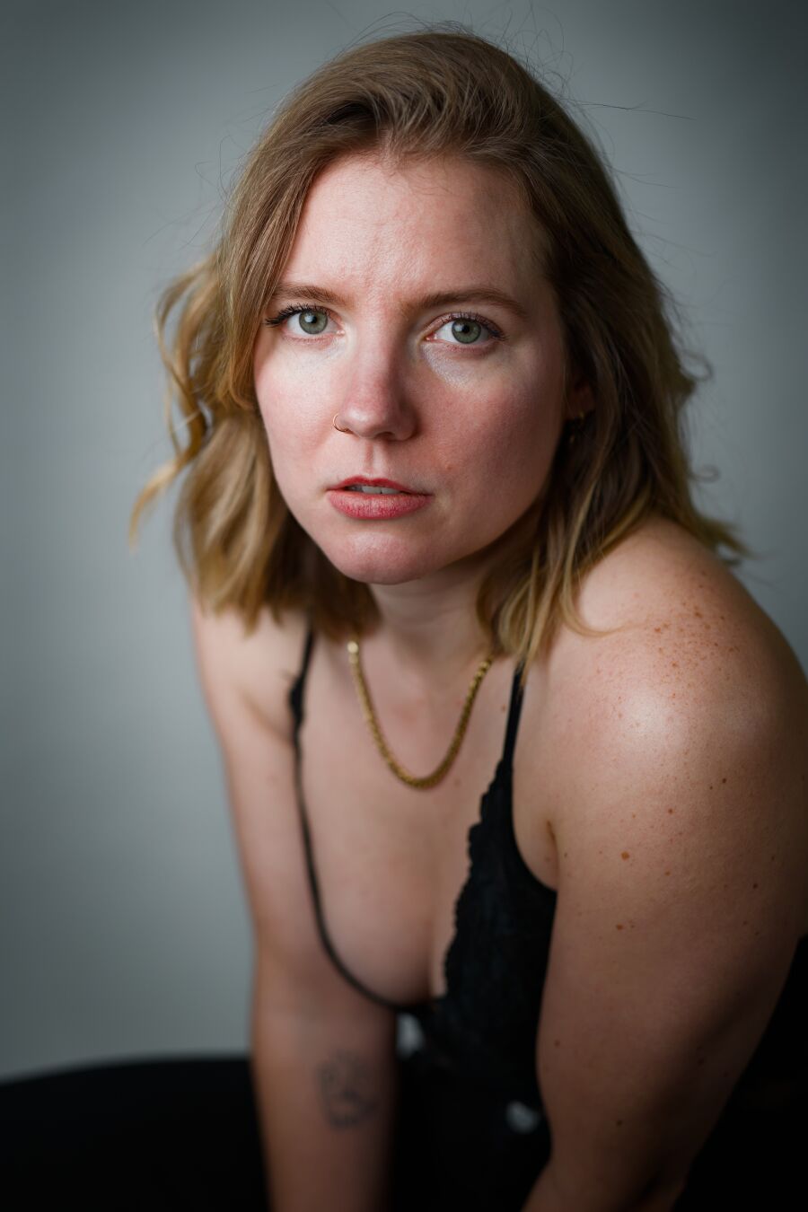 DE: Portrait einer blonden Frau mit grünen Augen. Sie trägt eine schwarze Hose, einen schwarzen BH und eine Goldkette. Sie sitzt mit dem Körper 90 Grad gedreht zur Kamera und dreht ihren Oberkörper zur Kamera. Ihr Blick geht direkt zur betrachtenden Person. Der Hintergrund ist verschwommen grau.

EN: Portrait of a blonde woman with green eyes. She is wearing black trousers, a black bra and a gold chain. She sits with her body turned 90 degrees to the camera and turns her upper body towards the camera. She looks directly at the person looking at her. The background is blurred gray.