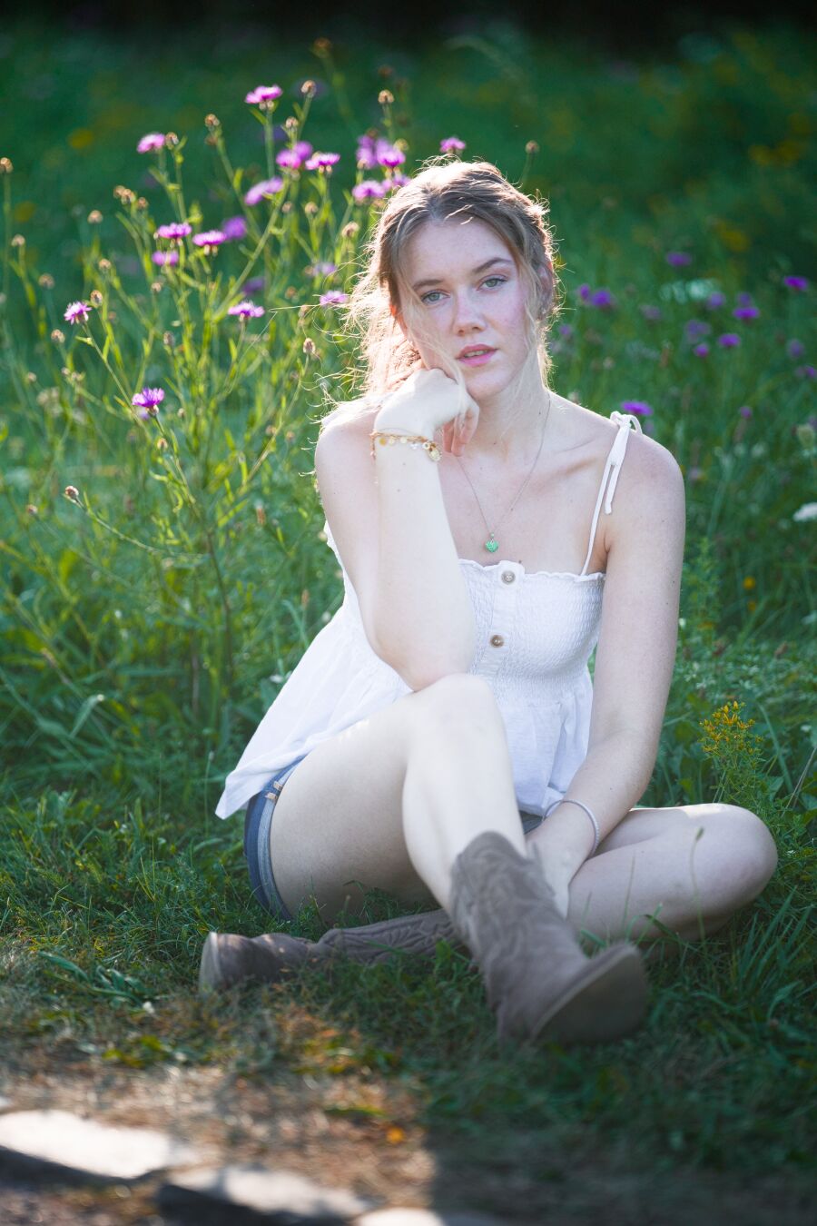 DE: Foto einer jungen blonden Frau mit grünen Augen. Sie trägt ein weißes Top, Jeans Hotpants und braune kurze Stiefel. Sie sitzt im Gras neben einem Weg. Der rechte Ellenbogen stützt gegen das rechte Bein und die angewinkelte Hand stützt den Kopf. Sie schaut mit leicht geöffnetem Mund direkt in die Kamera. Im Hintergrund sind lila Blüten zu erkennen, der Rest ist verschwommen grün.

EN: Photo of a young blonde woman with green eyes. She is wearing a white top, denim hot pants and brown ankle boots. She is sitting in the grass next to a path. Her right elbow is resting on her right leg and her bent hand is supporting her head. She is looking directly into the camera with her mouth slightly open. Purple flowers can be seen in the background, the rest is a blur of green.