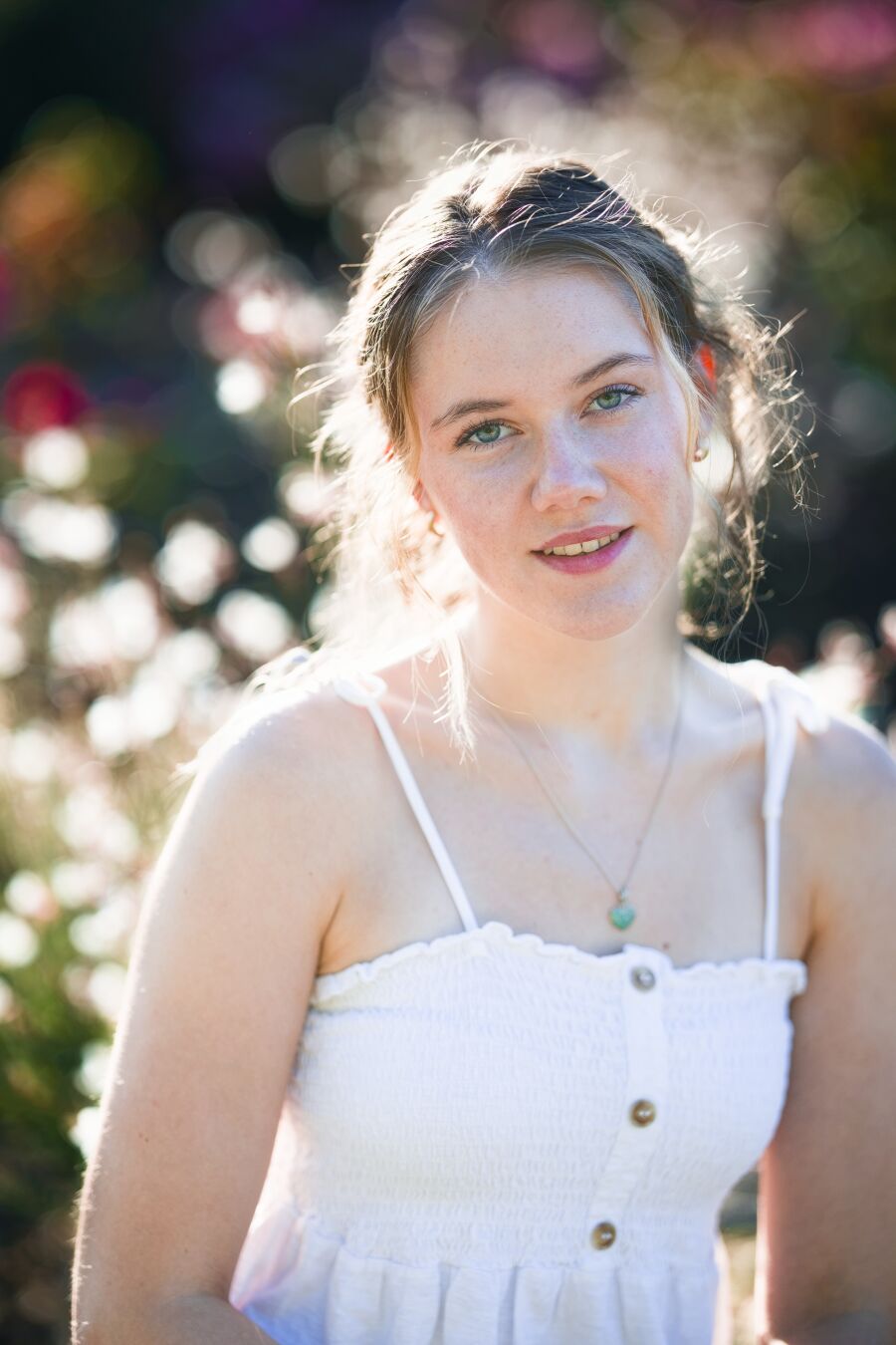 DE: Foto einer jungen blonden Frau mit grünen Augen. Sie trägt ein weißes Top. Sie schaut mit leicht lächelndem Mund direkt in die Kamera. Im Hintergrund sind Blüten zu erkennen, die warm im tief stehenden Sonnenlicht leuchten. Der Rest ist verschwommen bunt.

EN: Photo of a young blonde woman with green eyes. She is wearing a white top. She is looking directly into the camera with a slight smile on her lips. In the background, flowers can be seen glowing warmly in the low sunlight. The rest is blurred and colourful.