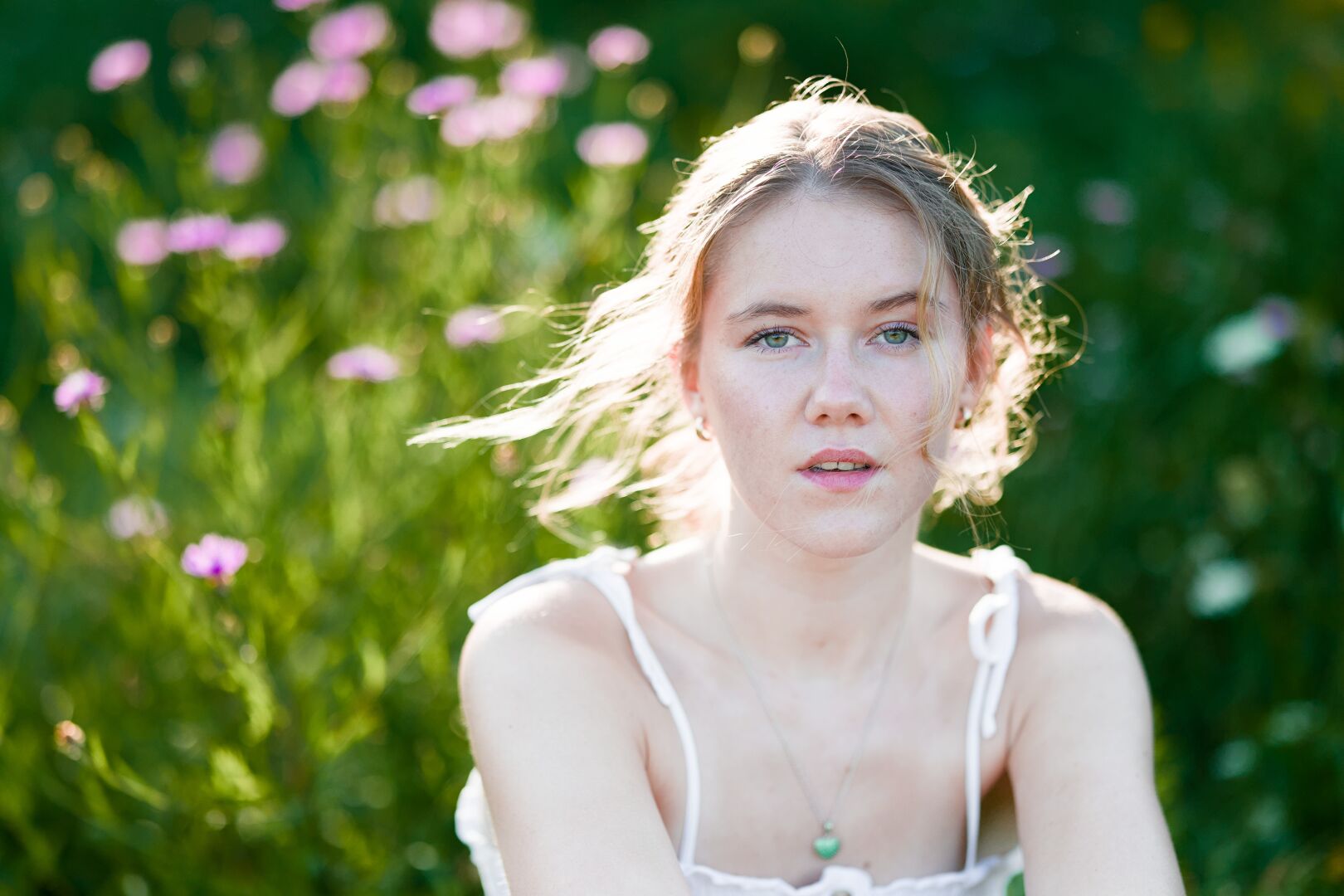 DE: Foto einer jungen blonden Frau mit grünen Augen. Sie trägt ein weißes Top. Sie schaut mit leicht geöffnetem Mund direkt in die Kamera. Die Haare werden  vom Wind verweht und leuchten warm von der tief stehenden Sonne. Der Hintergrund ist verschwommen grün und man links lila Blüten erkennen.

EN: Photo of a young blonde woman with green eyes. She is wearing a white top. She is looking directly into the camera with her mouth slightly open. Her hair is blowing in the wind and glowing warmly in the low sun. The background is blurred green and purple flowers can be seen on the left.