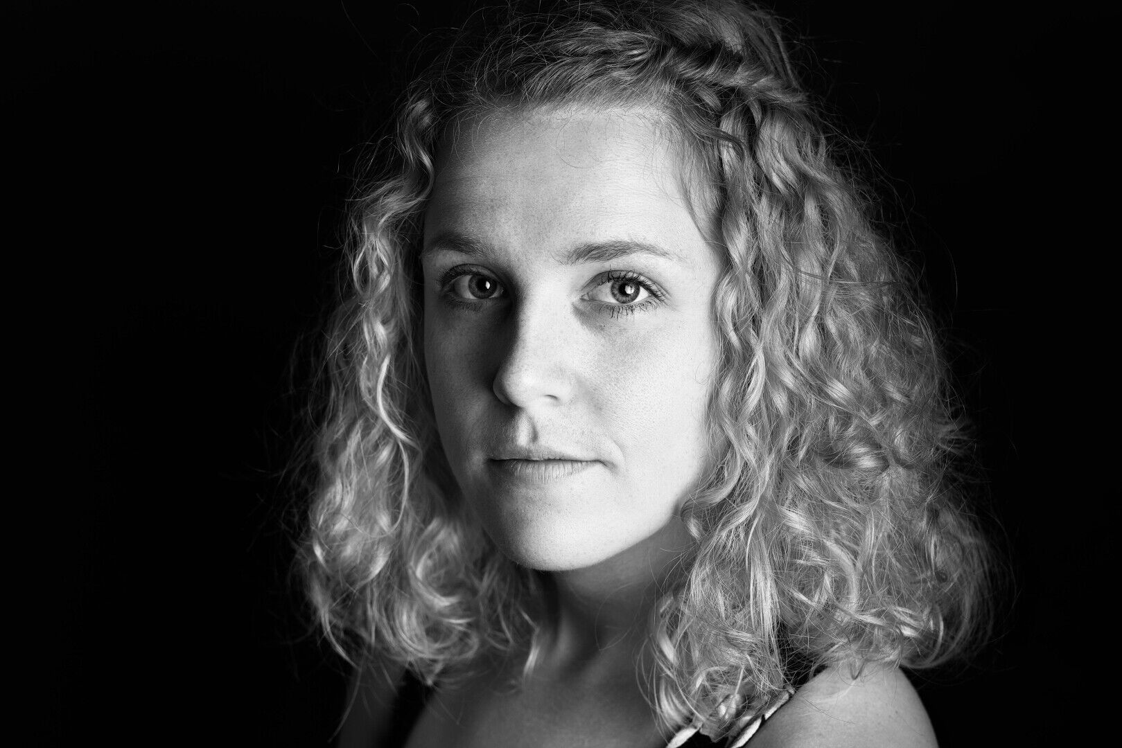 DE: Schwarz-weißes Bild eines blonden Mädchens mit Locken. Der Hintergrund ist komplett schwarz. Sie blickt nach links direkt in die Kamera.

EN: Black and white picture of a blond girl with curls. The background is completely black. She is looking to the left directly into the camera.